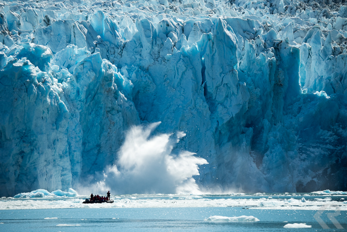 A glacier calves in front of a zodiac filled with people.