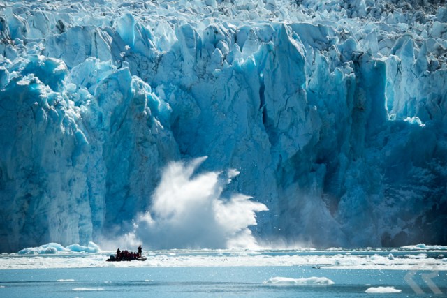 A glacier calves in front of a zodiac filled with people.